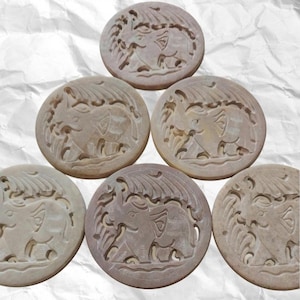 May include: Six beige, circular coasters with carved elephant designs. Each coaster shows a detailed relief of an elephant in a naturalistic setting. The coasters are displayed on a textured, white surface.