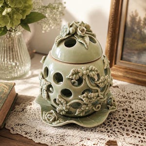 May include: A sage green ceramic oil burner with a removable lid, featuring a glossy crackle glaze finish. The spherical body is adorned with sculpted floral and leaf motifs and circular cutouts, resting on a scalloped, petal-shaped base.