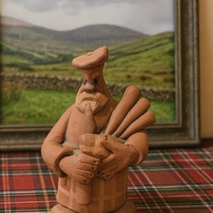 Puede incluir: Figura de terracota de un gaitero, con boina y sujetando una gaita. La figura tiene un rostro detallado y está sobre una superficie a cuadros. Una pintura de paisaje enmarcada está en el fondo.