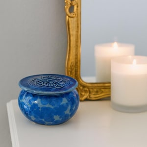 May include: A round, blue ceramic jar with a lid. The jar has a textured, cloud-like pattern in shades of blue. The lid features a detailed, embossed design. The jar sits on a white surface, with a gold-framed mirror and lit candles in the background.
