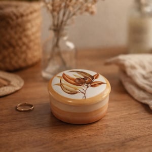 May include: A small, round ceramic trinket box with a botanical design in shades of brown and gold. The box has a cream-coloured top and a light orange base. A gold ring sits nearby on a wooden surface.