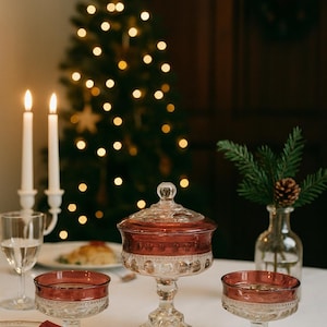 May include: A Christmas table setting with a covered glass candy dish with a claret red rim, two matching dessert dishes, and a lit Christmas tree in the background. Two lit candles and a glass of water are also visible.