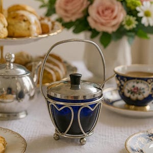 Könnte beinhalten: Eine silberfarbene Zuckerdose mit einem kobaltblauen Glaseinsatz und einem passenden Deckel. Die Schale hat ein herzförmiges Drahtdesign und einen kleinen Griff. Eine Tasse Tee, Gebäck und Blumen sind im Hintergrund.