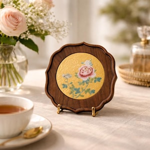 May include: A decorative wooden coaster with a gold centre featuring a floral design with pink and white flowers. The coaster has a scalloped edge and is displayed on a small brass stand.
