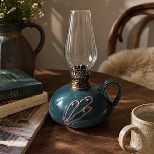 Könnte beinhalten: Eine türkisfarbene Öllampe aus Keramik mit einem transparenten Glaszylinder und Messingakzenten. Die Lampe hat einen Griff und ein rosa Blumendesign. Sie steht auf einem Holztisch mit einer Tasse und Büchern.