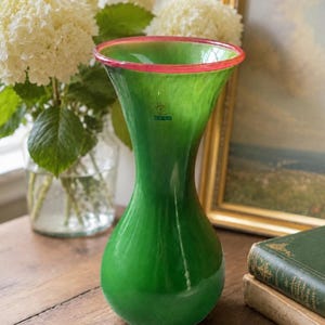 Può includere: Un vaso in vetro verde con un'apertura svasata e un sottile bordo rosso. Il vaso presenta una finitura liscia e lucida e un piccolo adesivo circolare blu e bianco con una grafica a spirale e del testo sul collo del corpo in vetro.