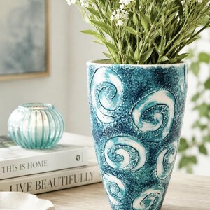 May include: A tapered ceramic vase featuring a textured, deep blue and teal surface with prominent white painted swirls. The vase is filled with a bouquet of small white flowers, showcasing a rough, handcrafted finish with visible colour variations.