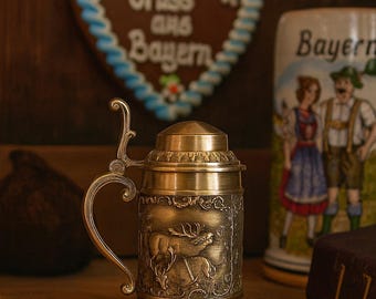 May include: A vintage brass beer stein with a detailed relief of a deer, featuring a hinged lid and ornate handle. The stein is set against a backdrop of Bavarian-themed decorations, including a heart-shaped gingerbread cookie and a ceramic mug.