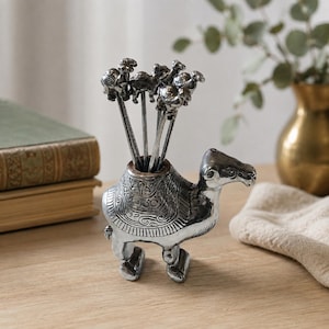 May include: A silver-toned metal camel-shaped holder containing five matching silver-toned metal picks. The camel figure features intricate embossed patterns on its back and sides, with a cylindrical opening at the top to store the slender picks.