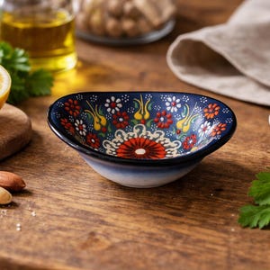 May include: A small, oval ceramic bowl with a dark blue exterior and a colourful floral pattern. The bowl is filled with a red and white design. The image also includes a lemon, nuts, and a small bowl of salt.