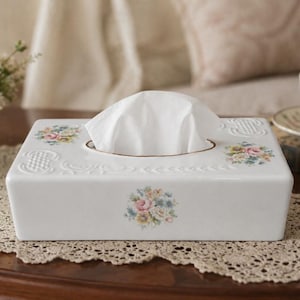 May include: A rectangular white ceramic tissue box cover featuring embossed textured patterns and delicate floral decals in shades of pink, yellow, and blue. The top surface has a central oval opening with a gold-toned rim, holding a white tissue.