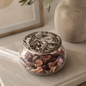 Pode incluir: Um pequeno frasco de vidro transparente cheio de flores secas e plantas, coberto por uma tampa decorativa prateada com desenhos de pássaros. O frasco está sobre um livro de tom neutro, com um vaso e uma obra de arte emoldurada no fundo.