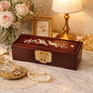 Puede incluir: Una caja de joyería rectangular de madera oscura con un cierre dorado. La tapa está decorada con una incrustación floral de flores blancas y hojas verdes. La caja está sobre una superficie de mármol, con una bandeja dorada y un collar de perlas cerca.