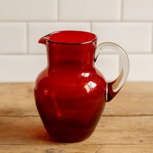 May include: A deep red glass jug with a clear handle. The jug has a rounded body and a spout. It is displayed on a wooden surface. This decorative jug is suitable for serving drinks.