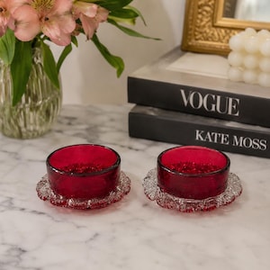 Peut inclure: Deux petits bols ronds en verre rouge profond avec des bases en verre transparent texturé présentant un bord festonné. Les bols ont un intérieur et un extérieur en verre rouge translucide lisse, reposant sur une base en verre transparent.