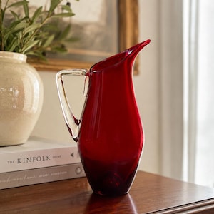 May include: A vibrant red glass jug featuring a smooth, rounded body and a clear, angular glass handle. The jug has a distinct pouring spout and a wide base, showcasing a translucent finish that catches the light, creating deep crimson tones.