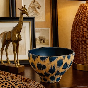 May include: A blue and gold leopard print bowl sits on a dark wooden surface. A gold giraffe sculpture and framed animal prints are in the background. A woven lamp and a leopard print runner complete the scene.