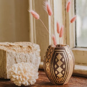 May include: A decorative vase with a brown and gold geometric pattern, holding pink dried flowers. A piece of white coral and a light-coloured stone are next to the vase, all on a wooden surface. The scene is lit by natural light.