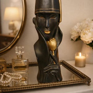 May include: A black and gold art deco bust sculpture of a woman holding a gold tulip. The sculpture has a hat and a long coat. The bust is displayed on a mirrored tray with a gold frame, alongside a perfume bottle and a candle.
