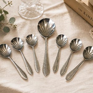 May include: A set of seven silver-toned metal spoons featuring a distinct shell-shaped bowl design with ribbed detailing. Each spoon handle displays a subtle etched leaf pattern, with one larger central spoon surrounded by six smaller matching spoons.