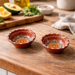 May include: Two small, round, terracotta bowls with detailed floral patterns in blue, yellow, and white. The bowls have a scalloped rim and are on a wooden surface, suitable for serving dips or sauces.