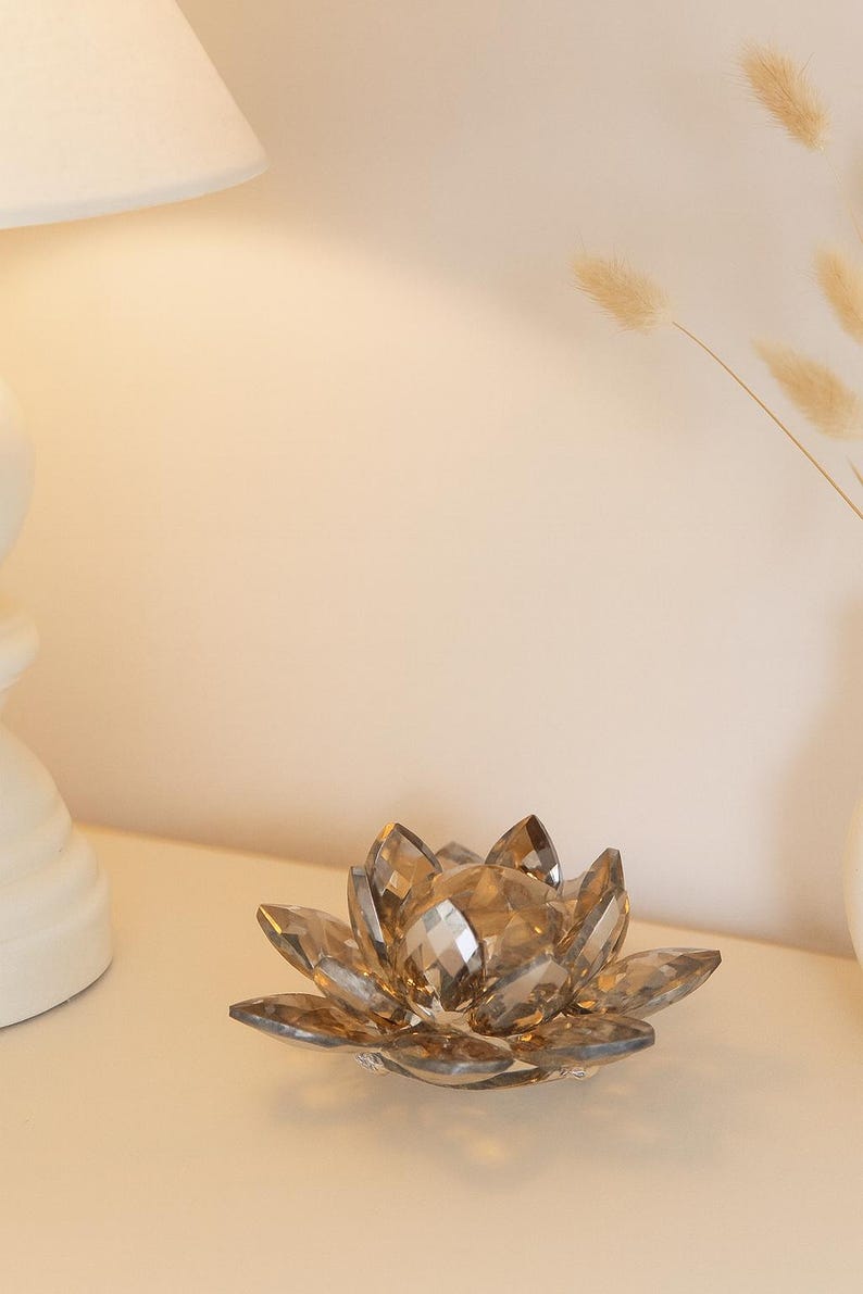 May include: A decorative crystal lotus flower with faceted petals. The flower is a warm amber color and sits on a white surface. The background includes a lamp and dried flowers.