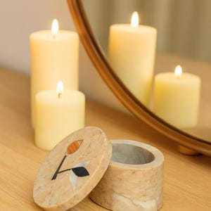 May include: A small, round marble box with a floral inlay on the lid. The box is open, revealing its interior. Several lit candles of different heights are in the background, with their reflections in a nearby mirror.