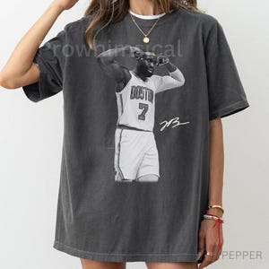 May include: Dark gray t-shirt featuring a black and white image of a basketball player in a white jersey with the word "BOSTON" and the number "7". The player is celebrating with arms raised. The shirt has a vintage, worn look.