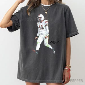 May include: Dark gray t-shirt featuring a full-body image of a football player in a white uniform with the number 11. The player is in action, with a signature on the right side. The shirt has a vintage, worn look.