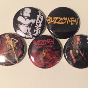 May include: Five round black and white buttons with the band name "Buzzov*en" printed in yellow and red. The buttons feature different images, including a baby, a person with long hair, and a red and black background.