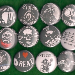 May include: A collection of 10 round, gray, button pins with various designs. The pins feature black and white illustrations of skulls, musical notes, a creature from the black lagoon, and other designs. The pins have white text on them, including "AVSKUM", "Disfem", "MOP 47", "NUCLEAR ATTACK", "O-BEAT", "DISCO", "UPP-U-DUPP-U", "Volfbrigad", "ANTI CIMEX", and "Country of Sweden".