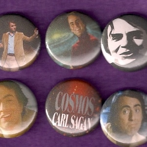 May include: Six round button pins featuring images of Carl Sagan.  The buttons are white with a silver rim.  One button features a black and white image of Carl Sagan's face.  Another button features a color image of Carl Sagan with a nebula in the background.  Another button features a color image of Carl Sagan with a galaxy in the background.  Another button features a color image of Carl Sagan with a planet in the background.  Another button features a color image of Carl Sagan with a star field in the background.  The last button features a black and white image of Carl Sagan with the text "Cosmos Carl Sagan" in white.