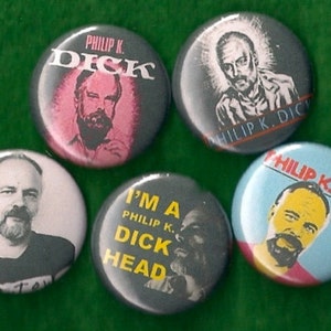 May include: Five round buttons with different images of Philip K. Dick. The buttons are black, white, blue, and gray with yellow, red, and pink accents. The text on the buttons reads "Philip K. Dick", "I'm a Philip K. Dick Head", and "Dick".