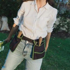 May include: A black gardening tool belt with green edging, worn around the waist. Two gardening tools are visible in the pockets. The person is wearing a white shirt and blue jeans. The background is a green lawn.