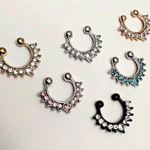 May include: An assortment of septum rings in various colors and styles. The rings feature a horseshoe design with decorative elements, including clear, pink, and blue gemstones. The rings are gold, silver, and black.
