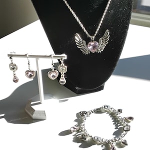 May include: A silver-toned jewelry set featuring a necklace with a heart pendant and wing accents, earrings, and a bracelet. The necklace and earrings are displayed on a black stand. The bracelet and earrings have pink heart-shaped and square-shaped accents.