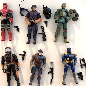 May include: Collection of six action figures, each in a combat-ready pose. The figures are in various colors, including red, black, blue, and green, with detailed armor and accessories like weapons and helmets. A variety of small black guns are scattered around the figures.