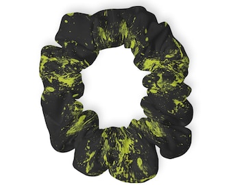 Könnte beinhalten: Ein kreisförmiges Haargummi aus Stoff mit schwarzer Grundfarbe und einem leuchtend neongelben Farbspritzermuster auf der Oberfläche. Das Accessoire weist durch das innenliegende Gummiband eine geraffte, wellige Textur auf.