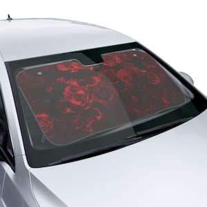 May include: A car windshield sun shade featuring a dense, repeating pattern of deep red roses with dark green leaves. The rectangular shade is positioned inside the front windshield of a white vehicle, covering the glass to block sunlight.