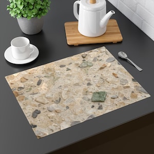 Pode incluir: Um lugar americano retangular com um padrão de granilite em tons de bege e castanho, com fragmentos de pedra incrustados em tons de cinzento, branco e verde. A superfície tem um aspeto salpicado e multicolorido com formas angulares.