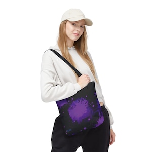 May include: Black tote bag with a vibrant purple splatter design. The bag is carried over the shoulder and features a solid black strap. The design includes a large central splash and smaller dots.