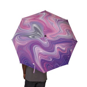 Puede incluir: Un paraguas abierto con un vibrante patrón de mármol en espiral en tonos morado intenso, lavanda claro, magenta y blanco. La cubierta muestra un diseño fluido similar a un líquido que irradia desde el centro, creando un efecto visual.