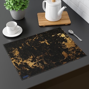 May include: Black placemat with gold abstract design. The placemat has a rectangular shape and is made of a textured material. The design features gold paint splatters and accents on a black background. A small text is visible in the bottom left corner.