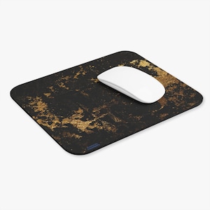 May include: A black rectangular mousepad with gold-colored paint splatter design. A white wireless mouse rests on the pad. The mousepad has rounded corners and a smooth surface. The design gives the mousepad a modern, artistic look.