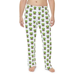 Op de afbeelding: Witte pyjamabroek met een herhalend patroon van een cartoonfiguur van groene broccoli met een lachend gezicht, een zonnebril op en een halter in de hand, vergezeld van de tekst "BROCCOLI PUMP" op een rood en zwart spandoek.