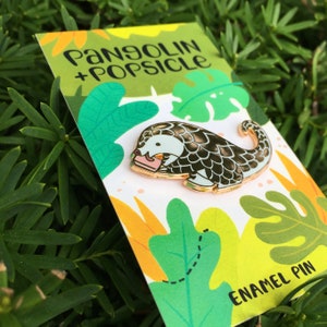Puede incluir: Un pin de esmalte dorado con un pangolín de dibujos animados que sostiene una paleta de helado rosa. El pin está en una tarjeta blanca con hojas tropicales verdes, amarillas y naranjas y el texto "Pangolin + Popsicle" y "Enamel Pin".