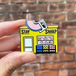 May include: A yellow enamel pin shaped like a storefront with the words "Stay Sharp" above the entrance. The storefront has a pink and blue window display and a pink pencil sharpener on top.