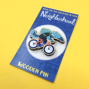 May include: Wooden pin featuring a blue and green anteater riding a red bicycle with blue wheels. The pin is on a blue card with the text "These are the creatures in your Neighbourhood" and "Wooden Pin".
