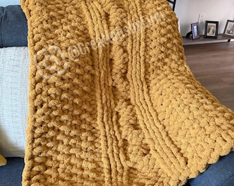 May include: A mustard yellow chunky knit blanket draped over a blue sofa. The blanket features a textured, cable-knit design and is made of thick, soft yarn. The blanket is a cozy home decor item.