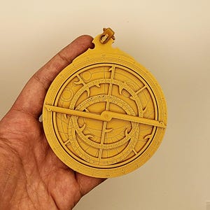 Könnte beinhalten: Ein goldfarbenes Astrolabium, ein historisches astronomisches Instrument, wird in einer Hand gehalten. Das Instrument weist komplizierte kreisförmige Designs auf, darunter Skalen und einen rotierenden Arm. Das Astrolabium ist ein Werkzeug für die Himmelsnavigation und Zeitmessung.